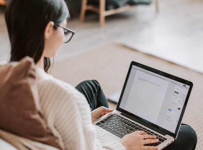 From above back view of unrecognizable female in casual wear typing on keyboard of laptop while working at home remotely